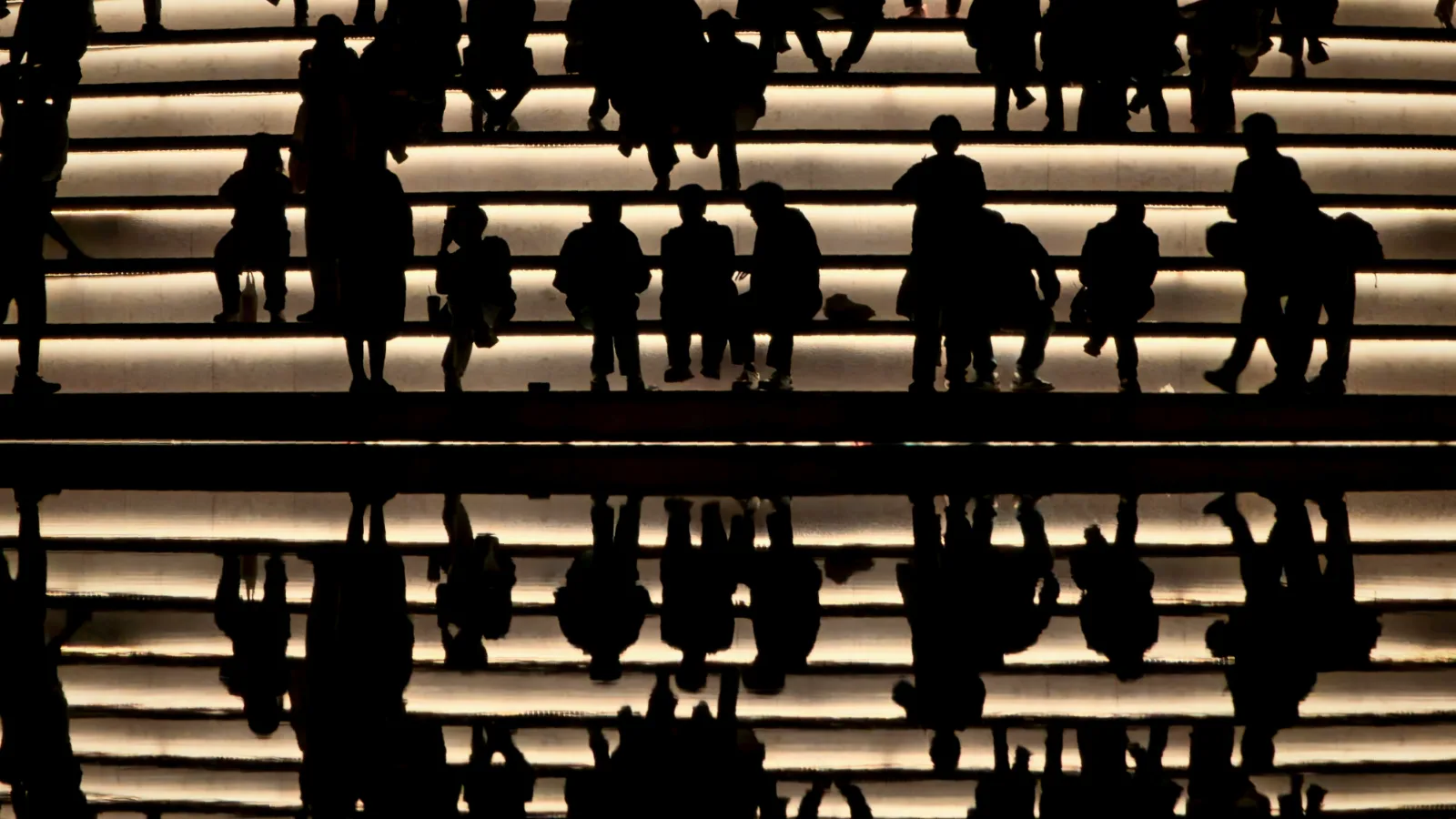 Silhouettes of people on brightly lit, rising steps, with their mirror image below.